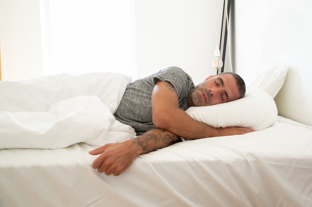 Adult man sleeping soundly on a white bed, tattoo visible, in a bright room.