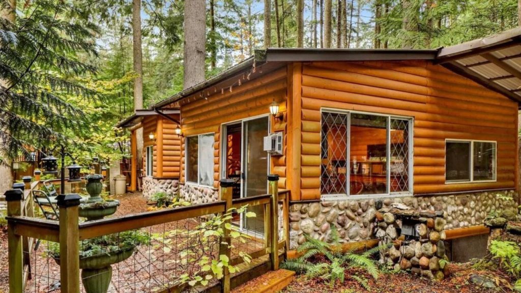A rustic log cabin surrounded by lush forest, featuring stone accents, large windows, and a cozy outdoor seating area.