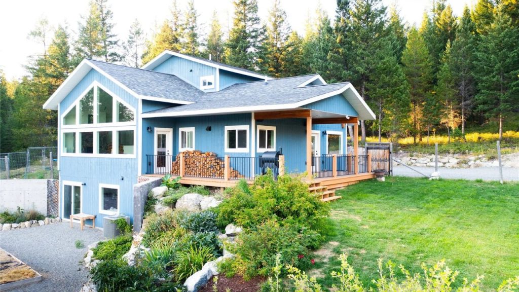 A blue mountain home with large windows, a covered porch, and lush greenery in Eureka, Montana.