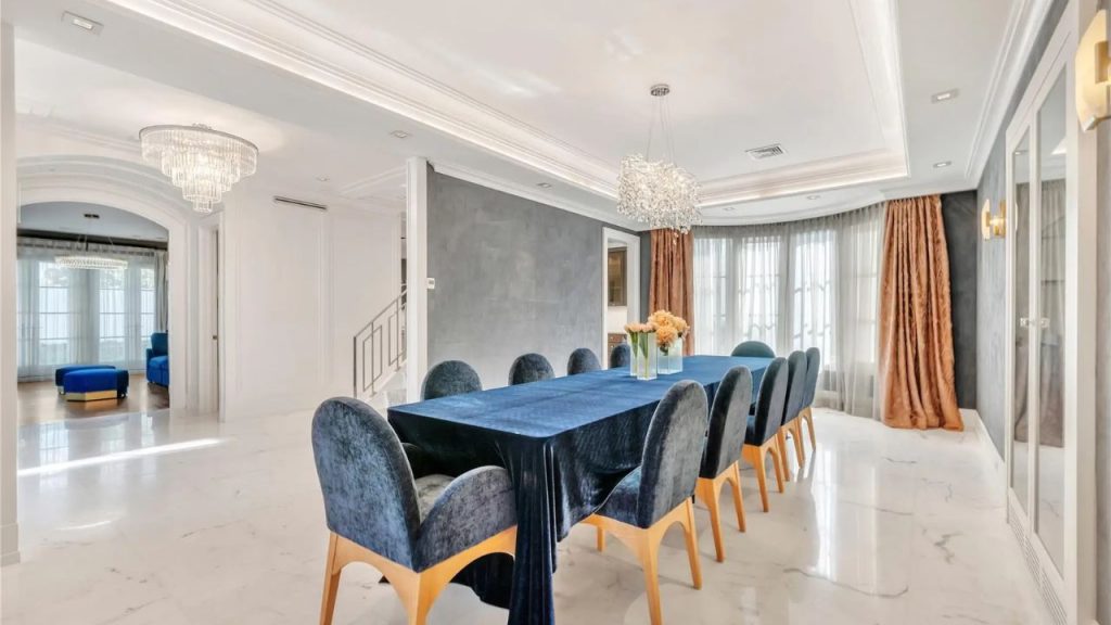 A luxurious dining room with a navy velvet table, plush chairs, and a crystal chandelier, framed by large windows and elegant drapery.