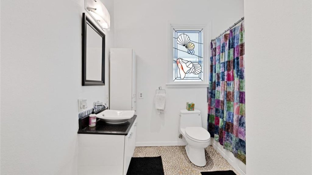 Bathroom with stained-glass window, pebble flooring, vessel sink, and colorful shower curtain.
