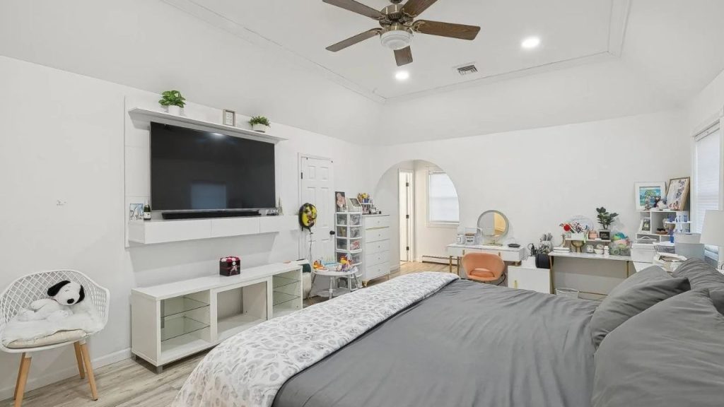 Spacious bedroom with neutral tones, a mounted TV, a cozy vanity area, and an arched doorway.