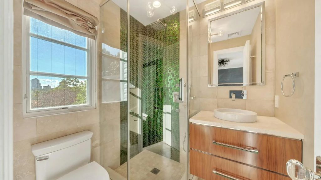 A modern bathroom with a glass-enclosed shower featuring green mosaic tiles, a wood-finish floating vanity, and a large window.