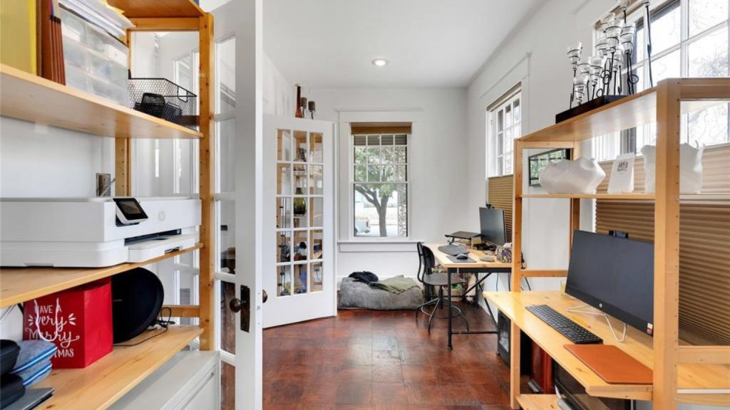Bright home office with wooden shelves, a dual-monitor desk, and large windows.