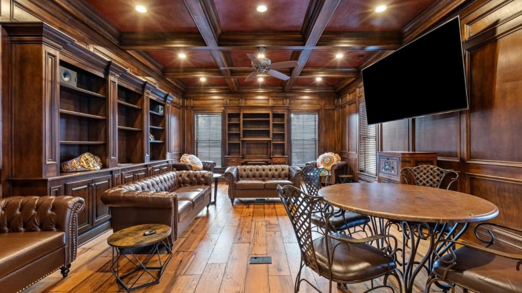 Wood-paneled library with leather sofas, bookshelves, and a mounted TV.