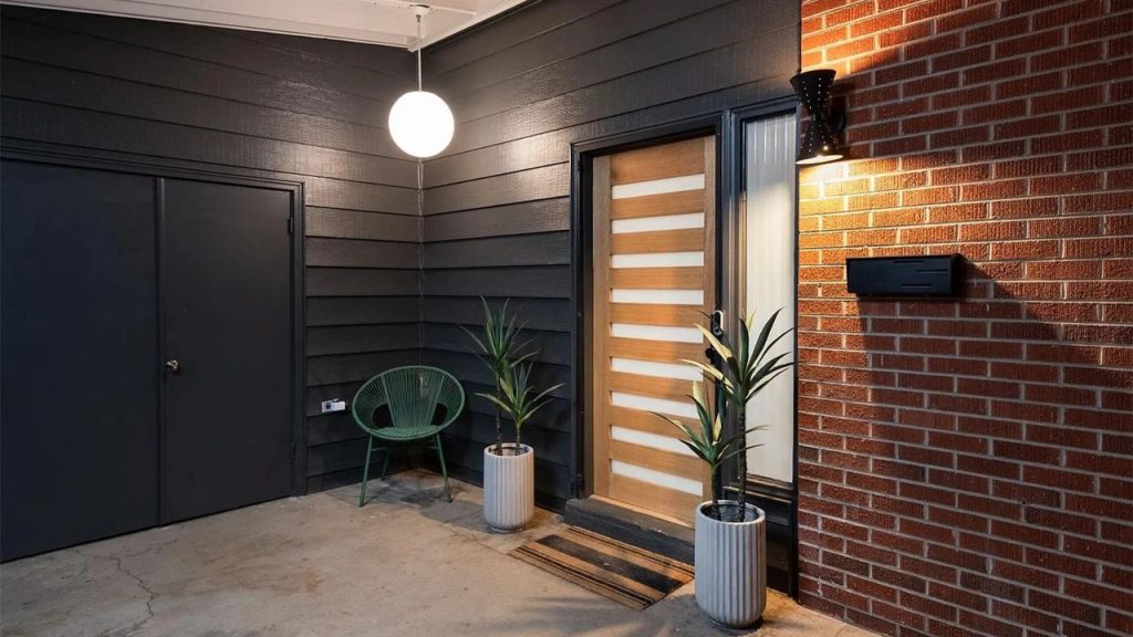 Stylish entryway with black siding, warm brick, a wooden door, and modern lighting.