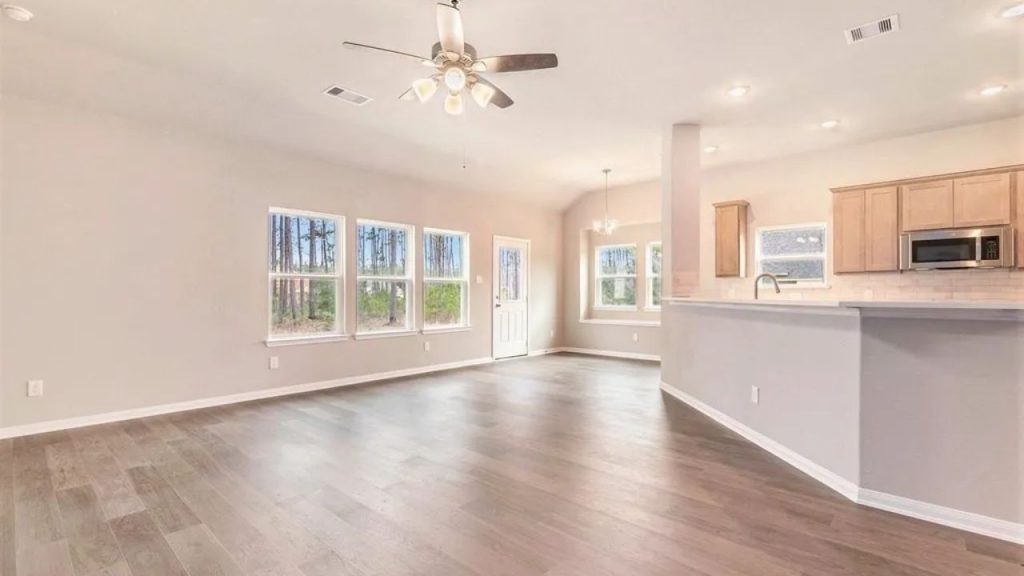 Bright open living area with large windows, ceiling fan, and an adjacent kitchen with wooden cabinets.