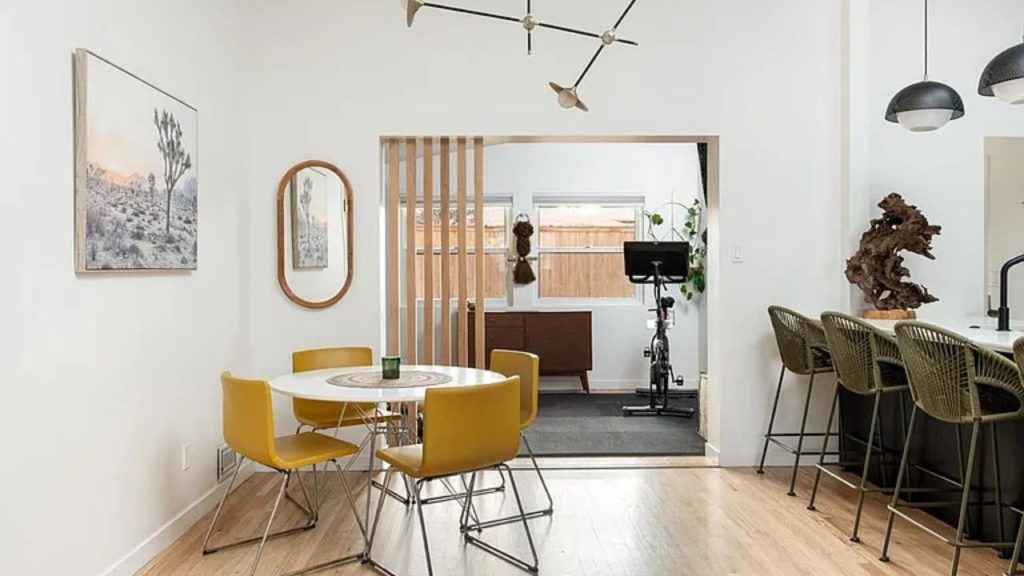 Mid-century dining area with mustard-yellow chairs, a round white table, and a wooden partition leading to a home gym.