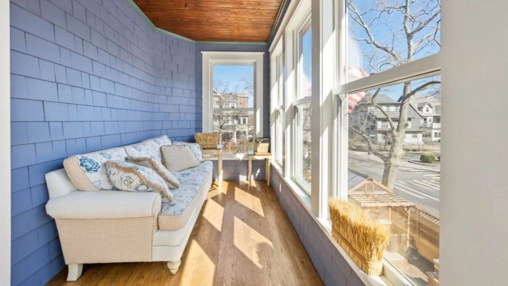 Sunlit cozy sunroom with blue shiplap walls, a cushioned sofa, and large windows overlooking a charming street.