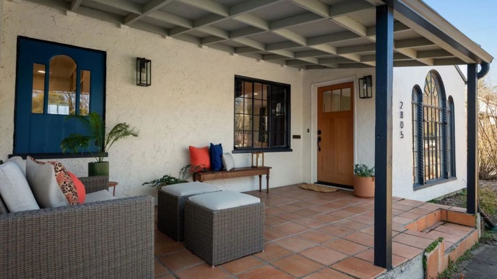 Mediterranean Revival front porch with terracotta tiles, wicker seating, wooden door, and blue accent window.
