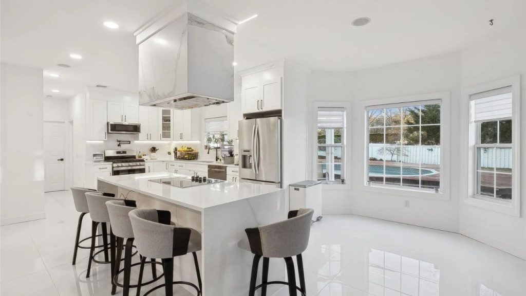 Elegant white kitchen with a large island, barstools, and a backyard view.