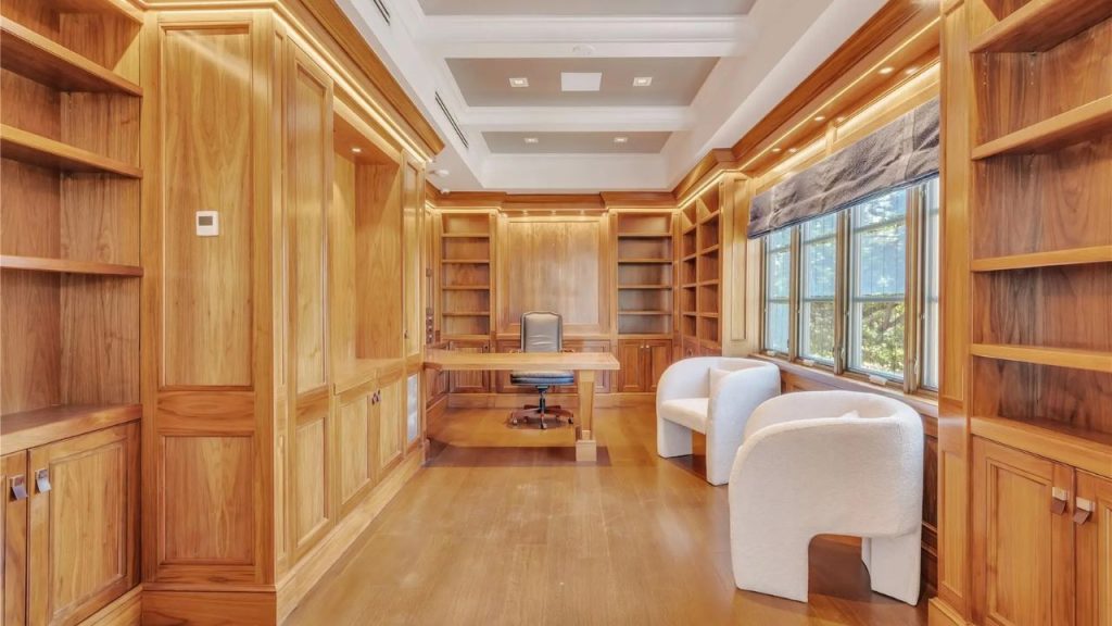A wood-paneled home office with built-in bookshelves, a central desk, and plush white chairs near large windows.
