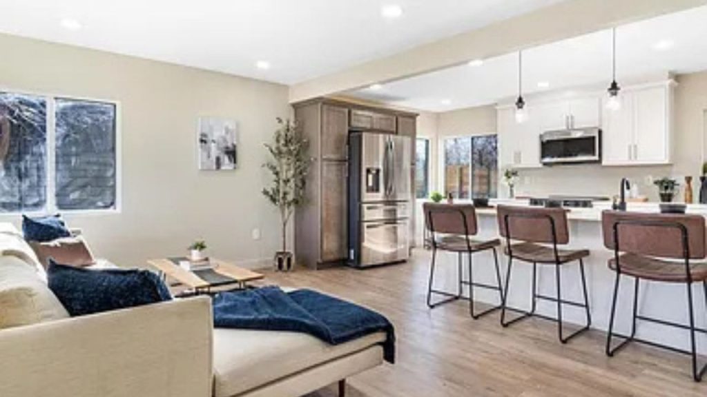 Open-concept living room and kitchen with a neutral sectional, wood flooring, and a stylish island with barstools.