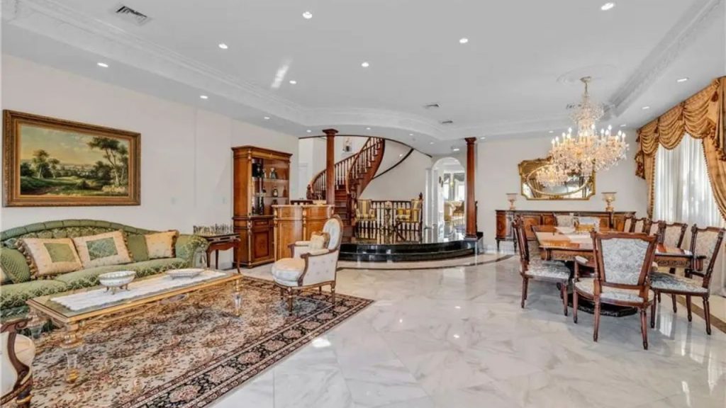 Elegant living and dining space with a curved staircase, chandeliers, marble flooring, and classic wooden furniture.