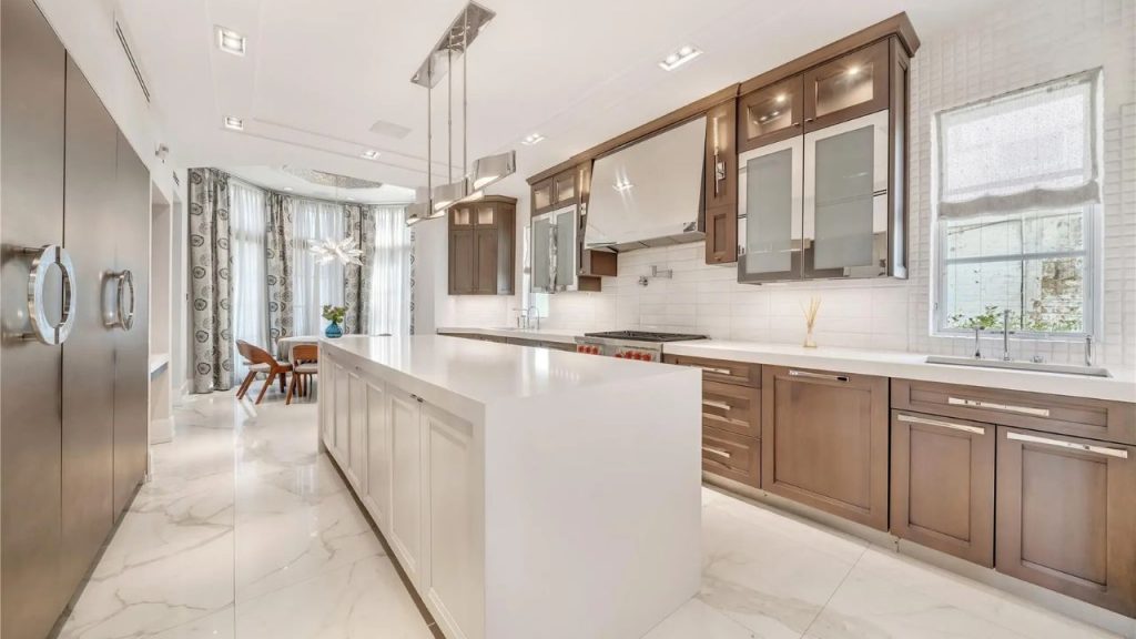 A bright, modern kitchen with a large white waterfall island, wood cabinetry, and a cozy dining nook with floor-to-ceiling windows.