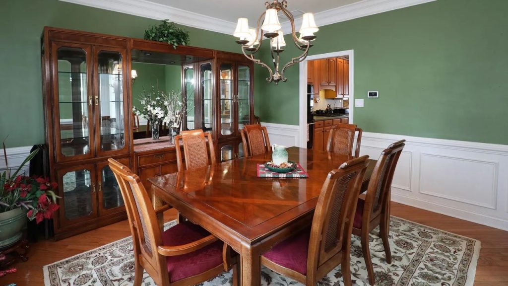 A warm dining room with a wooden table, cane-back chairs, and a china cabinet against green walls.