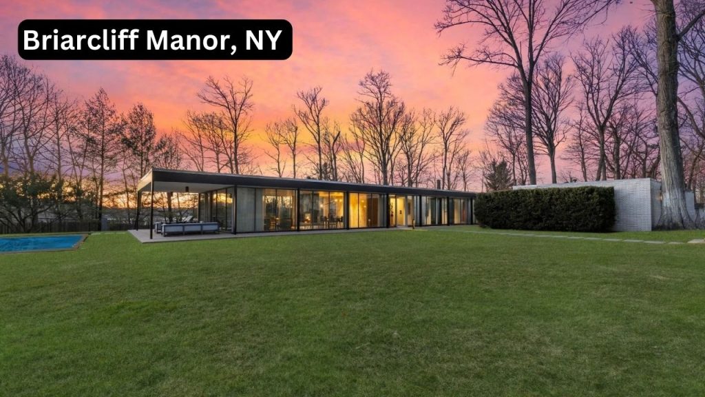 Modern glass-walled home in Briarcliff Manor, NY at sunset with large lawn and pool.
