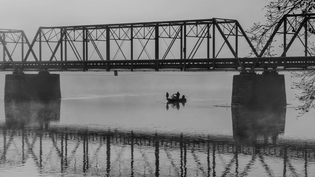 grayscale photo of person riding on horse on river near bridge