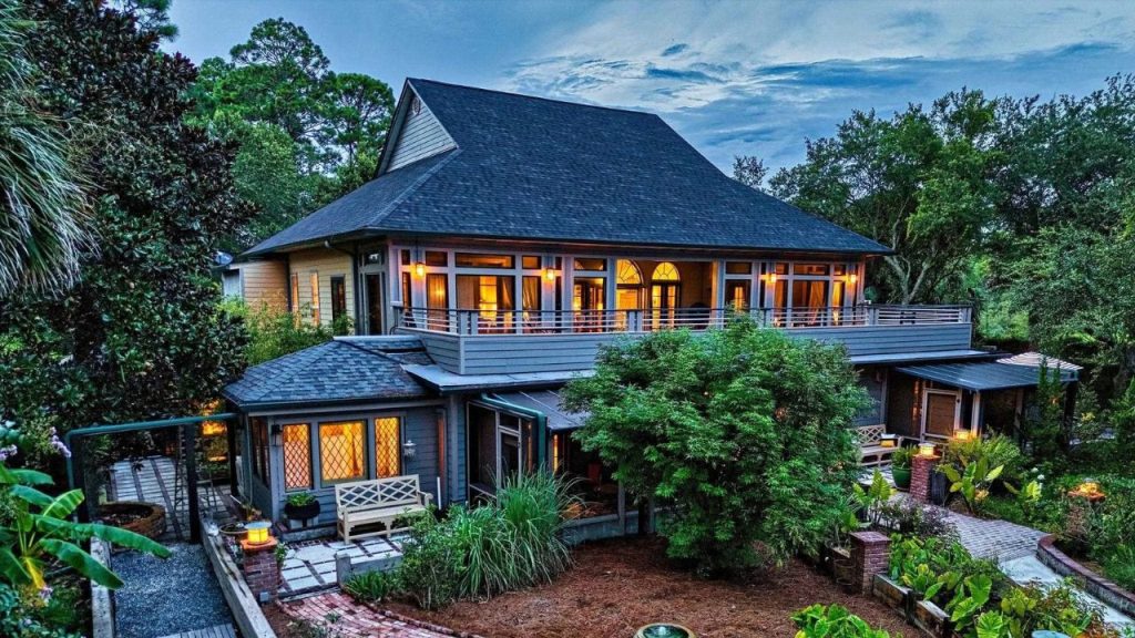 Colonial-style home at dusk with glowing windows, wraparound porch, and tropical landscaping.