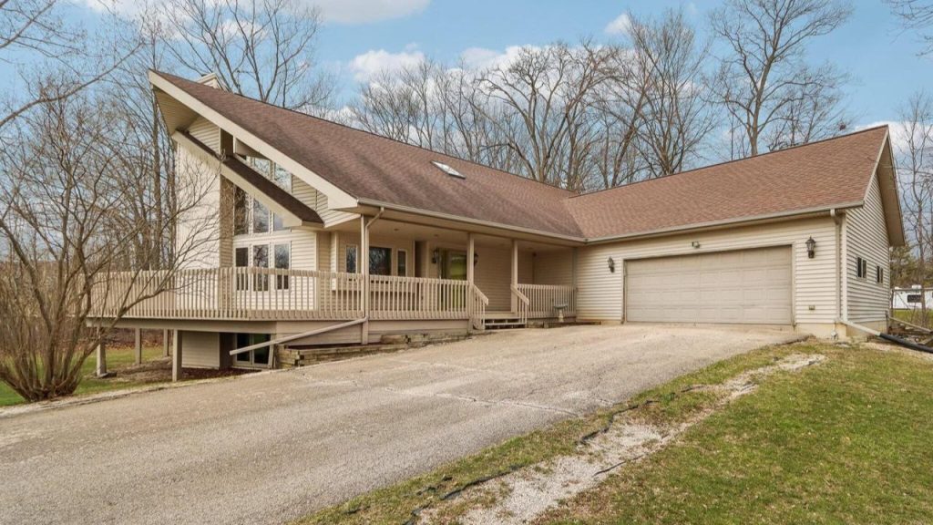 A charming colonial-modern house with a steep roof, large windows, a welcoming porch, and a garage, nestled among bare trees and a gravel driveway.