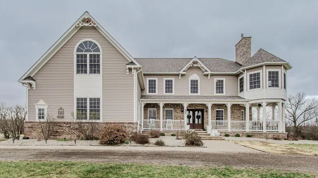 A charming Colonial Modern home with beige siding, a wraparound porch, and large windows, set against a green lawn.
