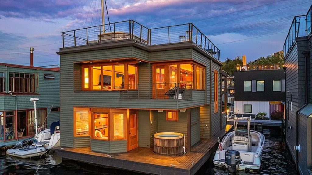 Modern two-story floating home with rooftop deck and hot tub, warmly lit at dusk.