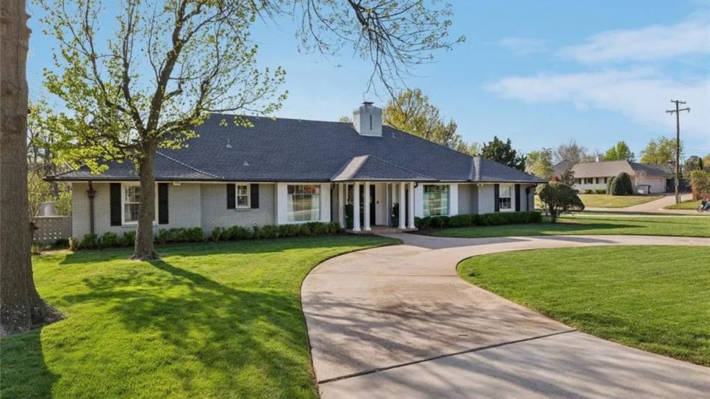 A charming Colonial Modern home with a gray roof, white brick walls, and a curved driveway surrounded by green lawns and trees.