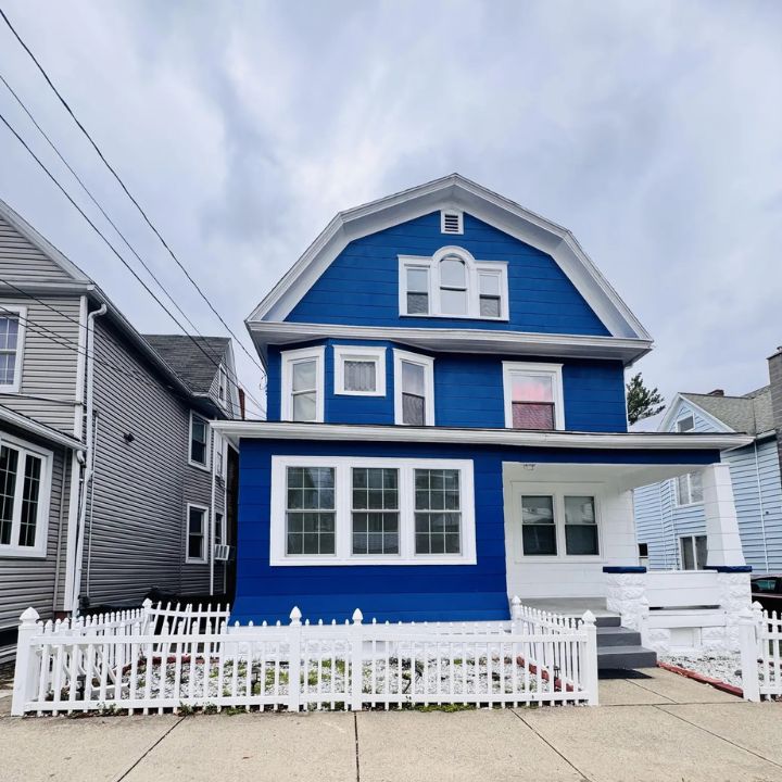 A vibrant blue colonial home with a white picket fence and porch, nestled among gray neighbors under a cloudy sky.