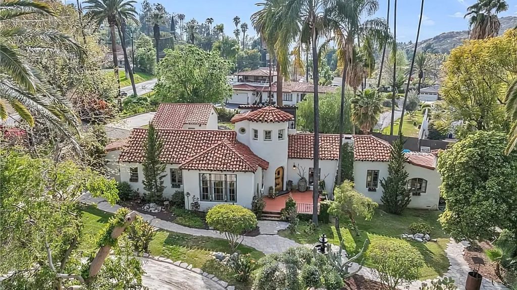 A stunning Spanish Colonial home with a red-tiled roof and white stucco walls, nestled among palm trees and greenery on a sunny hillside.
