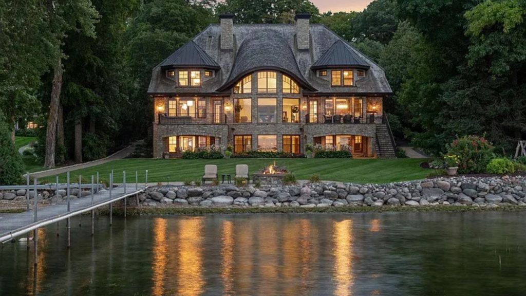 A luxurious stone home by a tranquil lake, featuring large windows, a fire pit, and a dock, surrounded by trees at dusk.
