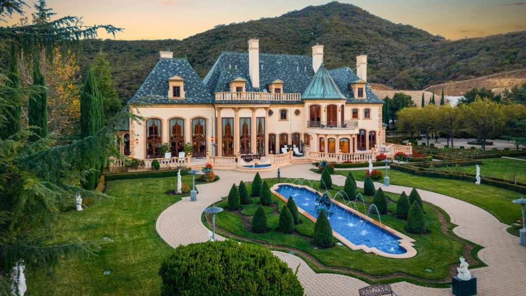 A luxurious colonial-style mansion with a slate roof, large arched windows, and a manicured garden featuring a blue fountain, set against a hilly backdrop.