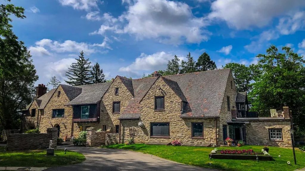 A beautiful stone house with steep roofs and chimneys, surrounded by trees and a colorful garden under a bright sky.