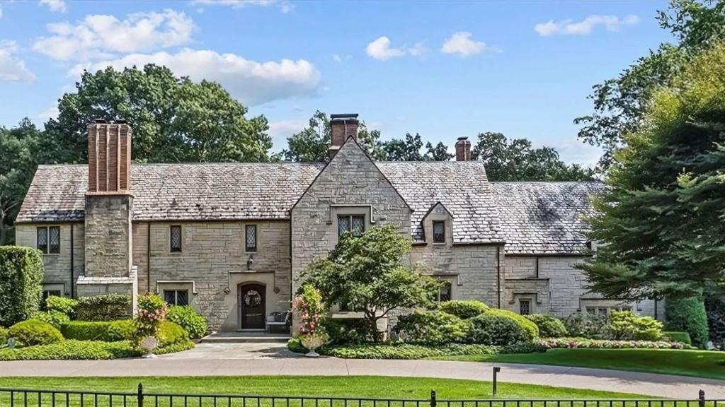 A beautiful colonial-style home with a stone exterior, slate roof, and lush gardens, framed by mature trees under a bright sky.