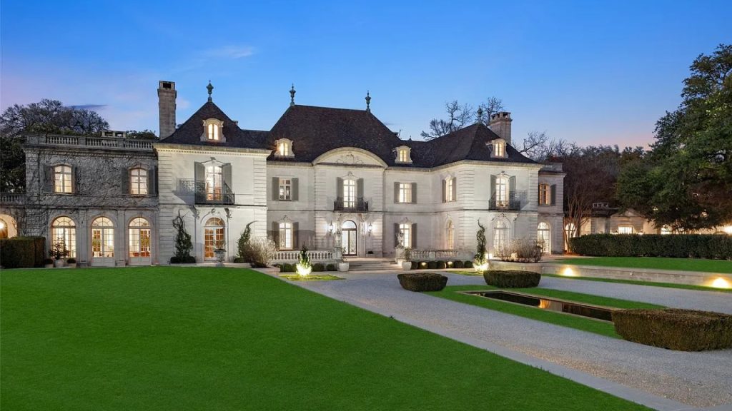 A grand Colonial modern home with a symmetrical facade, lit windows, and a lush lawn at dusk.