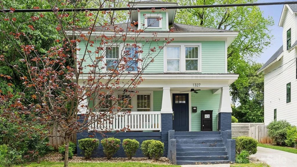 A charming two-story home with a mint green exterior, navy base, and white columns, surrounded by trees and bushes.
