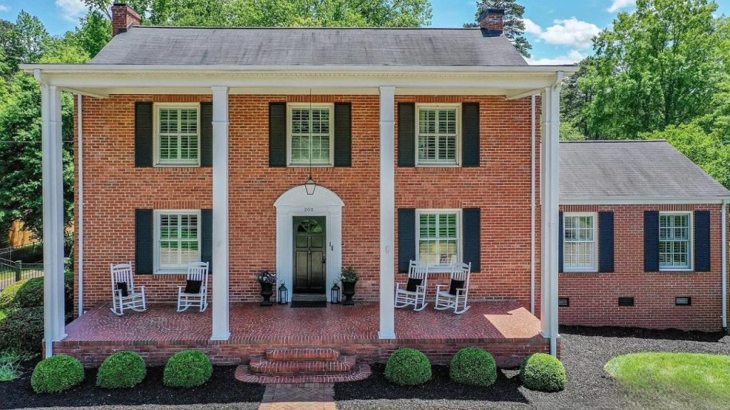 A charming red brick Colonial home with white columns, a welcoming porch with rocking chairs, and lush greenery under a bright sky.