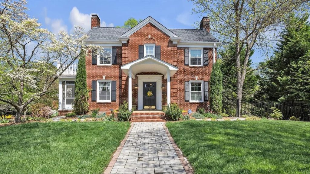 A charming Colonial-style home with red brick, white trim, and a stone pathway leading to a porticoed entrance, surrounded by green lawn and blooming trees.