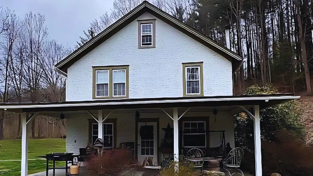 A charming colonial home with a white facade, green-trimmed windows, and a covered porch, set against a wooded backdrop.