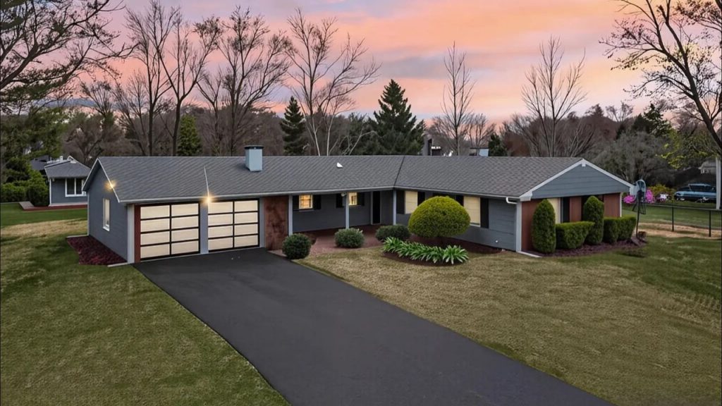 A charming Colonial Modern home with a gray exterior, double garage, and manicured lawn, framed by trees under a colorful sunset.