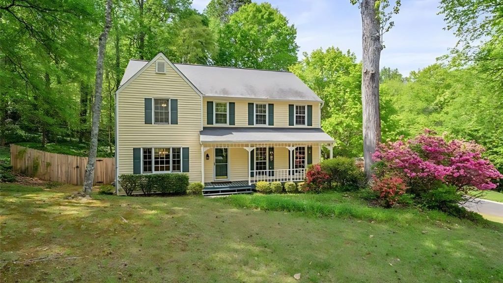 A charming yellow colonial home with green shutters, a front porch, and pink azaleas, nestled among tall trees and a green lawn.