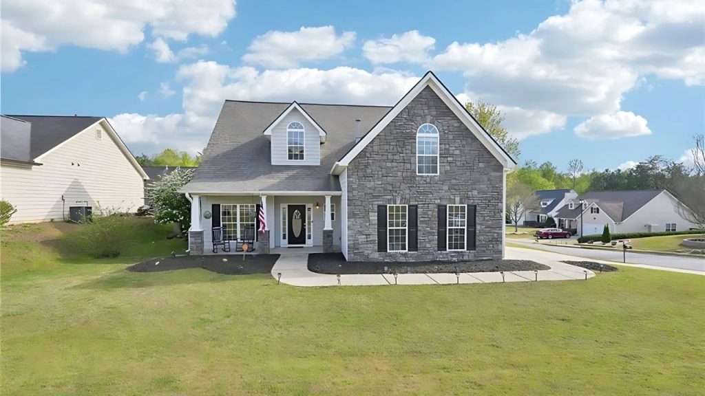 A charming colonial-style home with a stone facade, front porch, and lush green lawn under a bright, partly cloudy sky.
