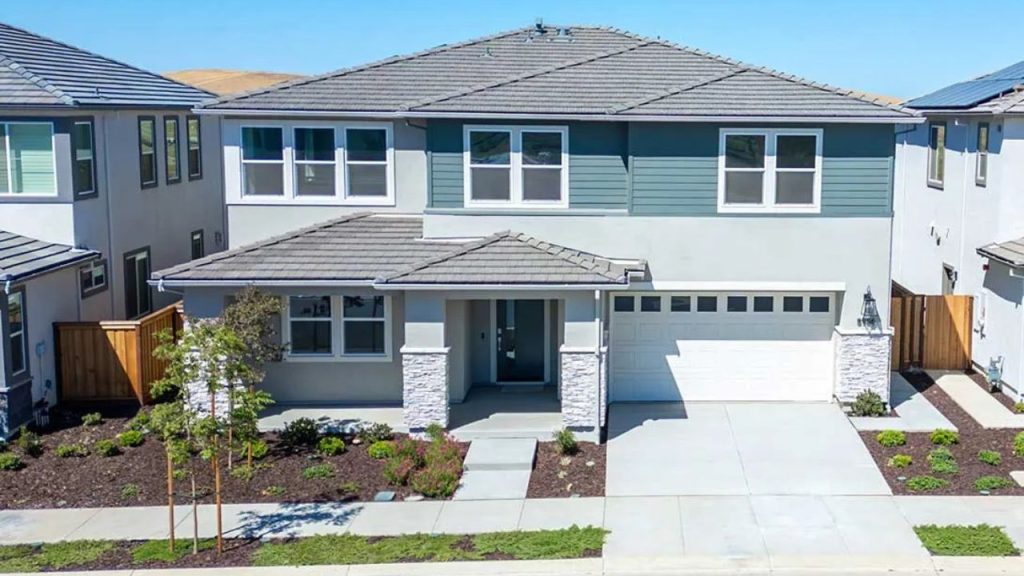 A modern two-story suburban home with a gray and teal exterior, stone pillars at the entrance, a double garage, and a tidy front yard with young trees under a clear blue sky.