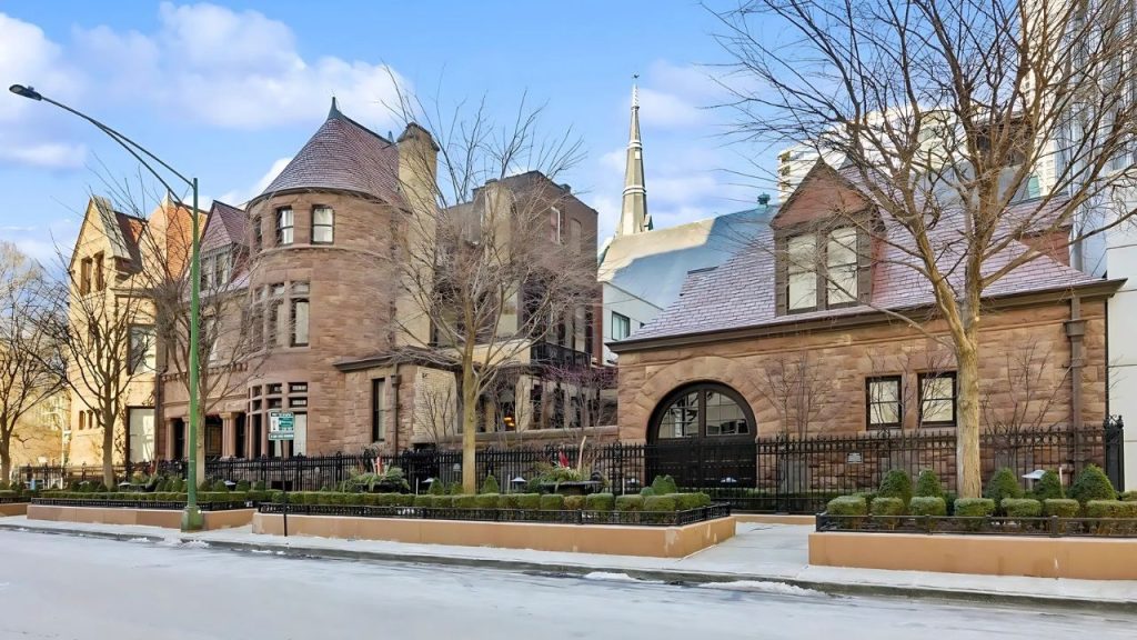 A historic brownstone with turreted corners and arched windows sits on a snowy street, surrounded by bare trees and a wrought-iron fence, with a church spire in the background.