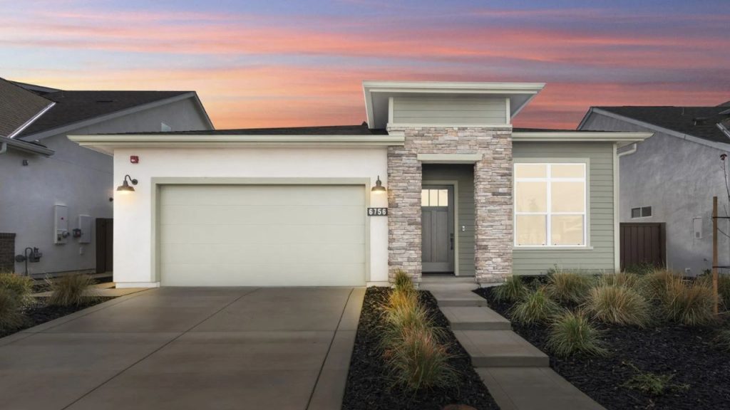 A modern coastal home with a stone and siding facade, featuring a large front window and a paved walkway with grasses, set against a colorful sunset sky.