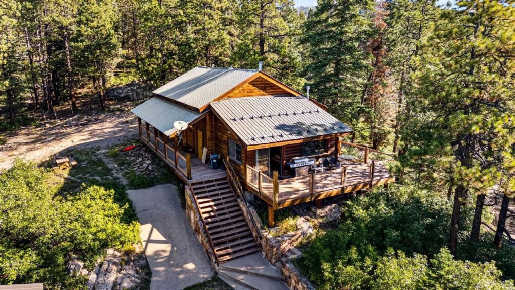 Cabin surrounded by forest with a wraparound deck and metal roof.