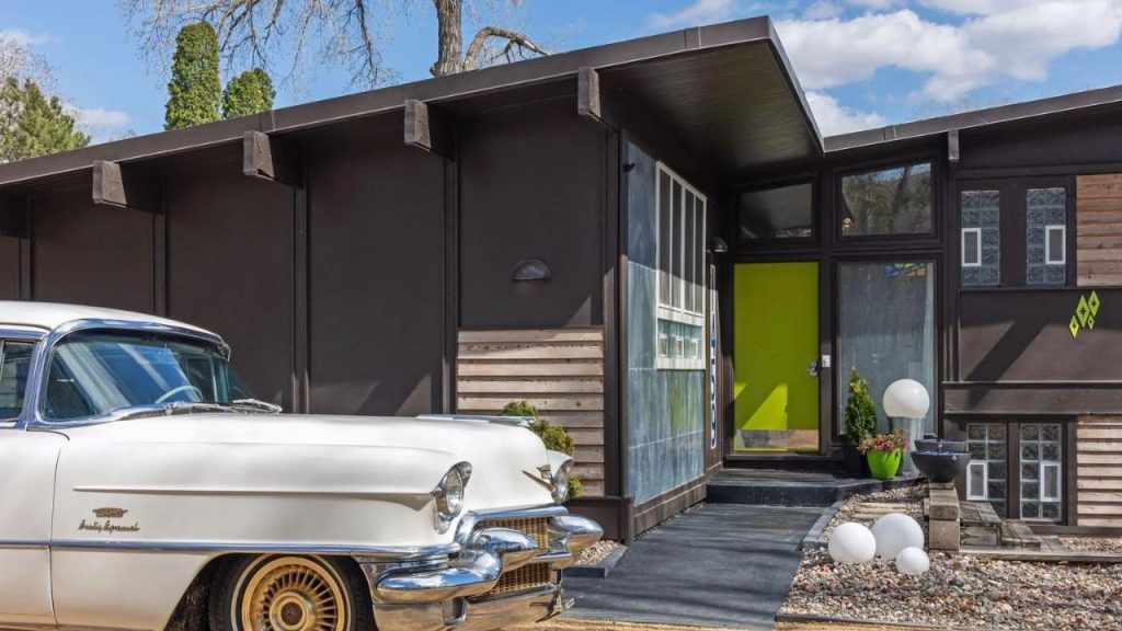 Midcentury home with lime green door, dark siding, and a vintage white Cadillac parked outside.