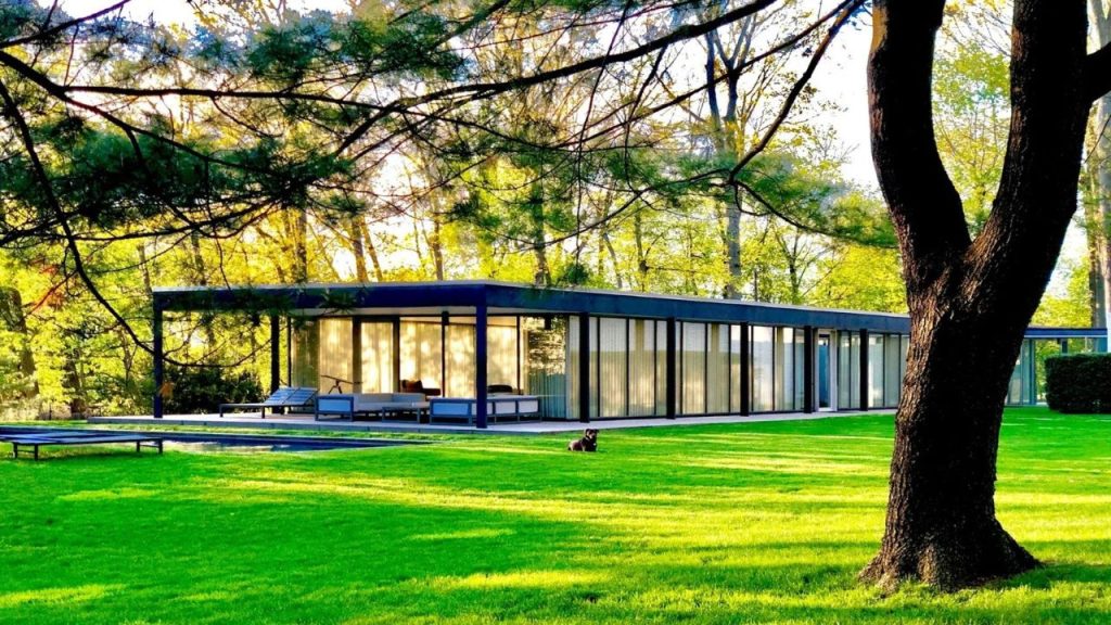 Modern glass house nestled in greenery with lounge chairs and a dog resting on the lawn.