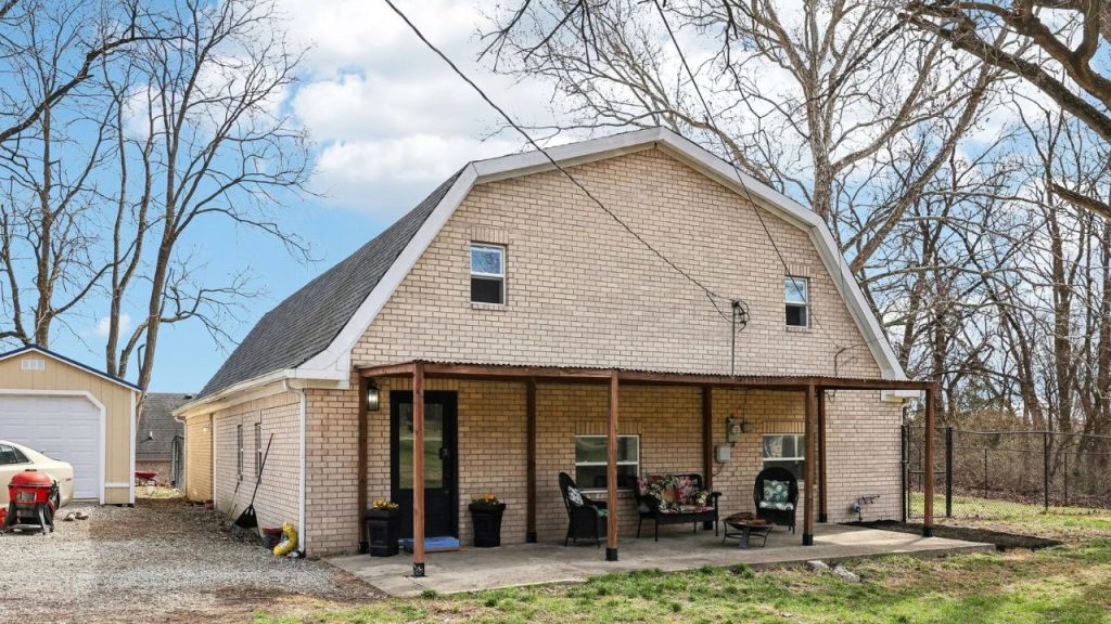 Gambrel-roof brick house with front porch and outdoor seating in a wooded yard.