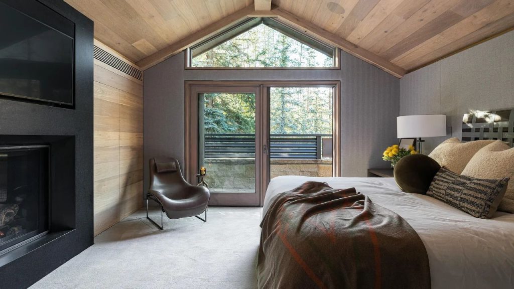 A cozy bedroom with a plush bed, fireplace, and forest view through large windows.
