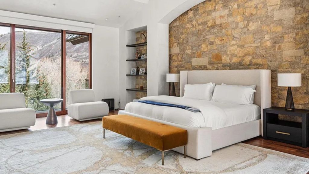 A cozy bedroom with a stone wall, white bed, orange bench, and large windows showcasing mountain views, plus armchairs and lamps.